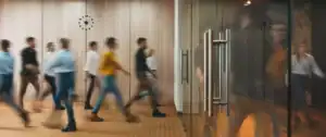 Motion-blurred people walking past a wood-paneled wall with a clock in a modern office hallway.