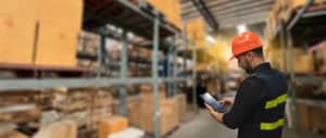 Worker in hard hat using tablet in a large, blurred warehouse setting.