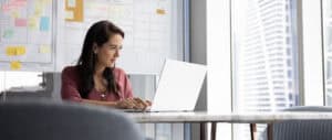 A woman smiles while typing on her laptop at a desk in a modern high-rise office.