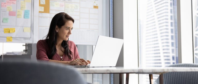 A woman smiles while typing on her laptop at a desk in a modern high-rise office.