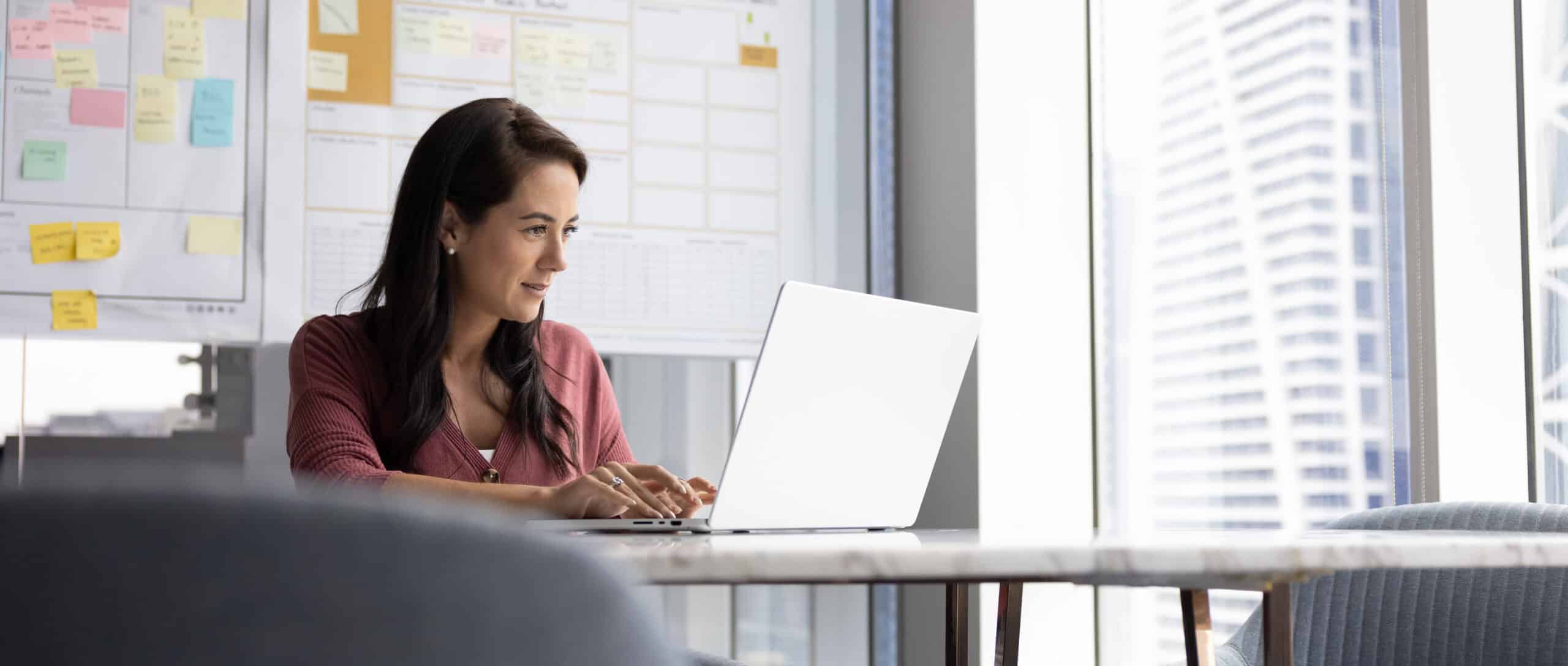 A woman smiles while typing on her laptop at a desk in a modern high-rise office.