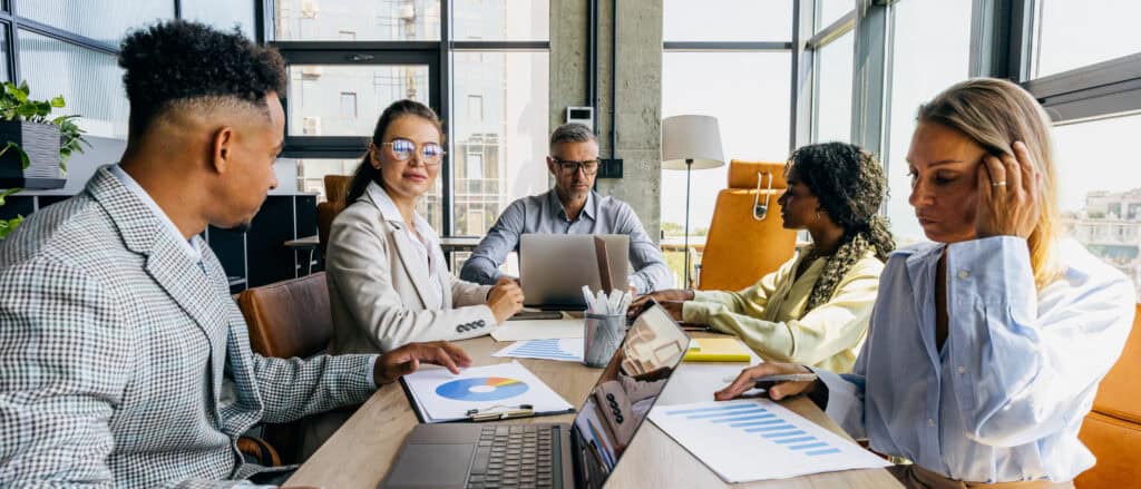 A diverse team of professionals analyzes charts and laptops around a conference table in an office.