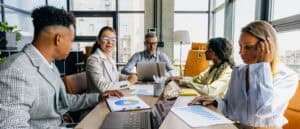 A diverse team of professionals analyzes charts and laptops around a conference table in an office.