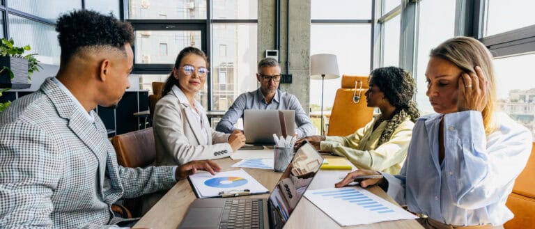A diverse team of professionals analyzes charts and laptops around a conference table in an office.