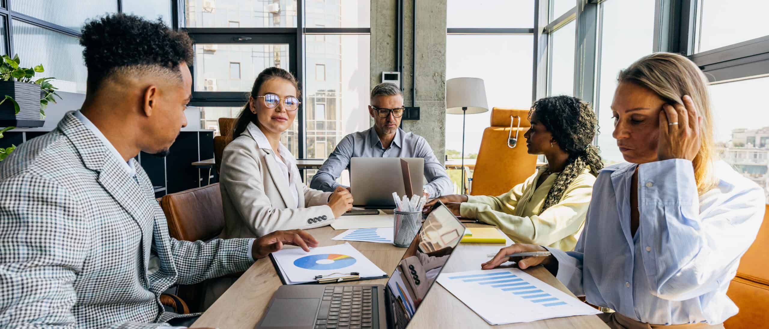 A diverse team of professionals analyzes charts and laptops around a conference table in an office.