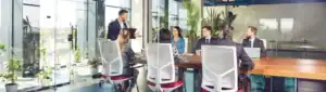 Professionals in business attire having a meeting around a long wooden table in a glass office.