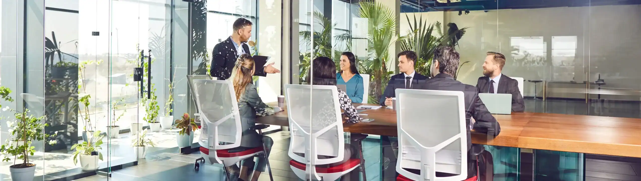 Professionals in business attire having a meeting around a long wooden table in a glass office.